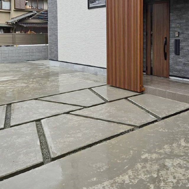 幾何学的なデザインのコンクリート舗装と、木目調の縦格子スクリーンがある玄関前のスペース